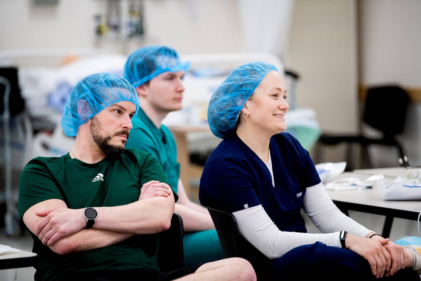 Three individuals wearing blue surgical caps sit attentively in a clinical setting, with one smiling and the others focused.